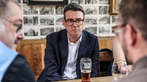 Andy Burnham, Mayor of Greater Manchester, in a dark suit and glasses, sitting at a table with a beer.
