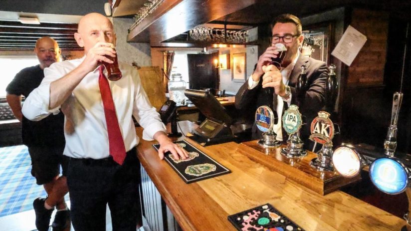 Two men drinking beer at a pub bar with several beer taps and an empty glass rack above.