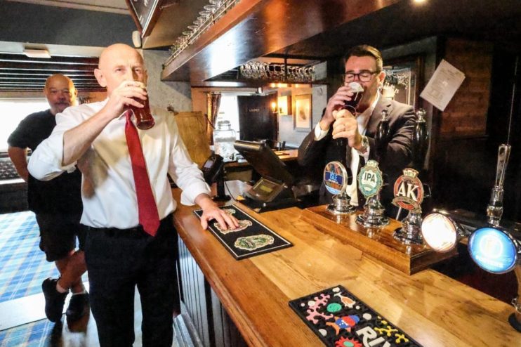 Two men drinking beer at a pub bar with several beer taps and an empty glass rack above.