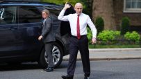A man in a white shirt and red tie carries his jacket, walking away from a dark SUV with a woman in the background.