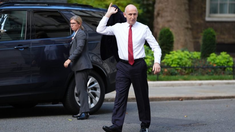 A man in a white shirt and red tie carries his jacket, walking away from a dark SUV with a woman in the background.