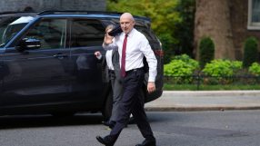 Bald man in white shirt and red tie walking past a dark SUV, holding a jacket over his shoulder