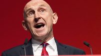 A bald man in a dark suit and red tie speaks passionately at a podium against a red background.
