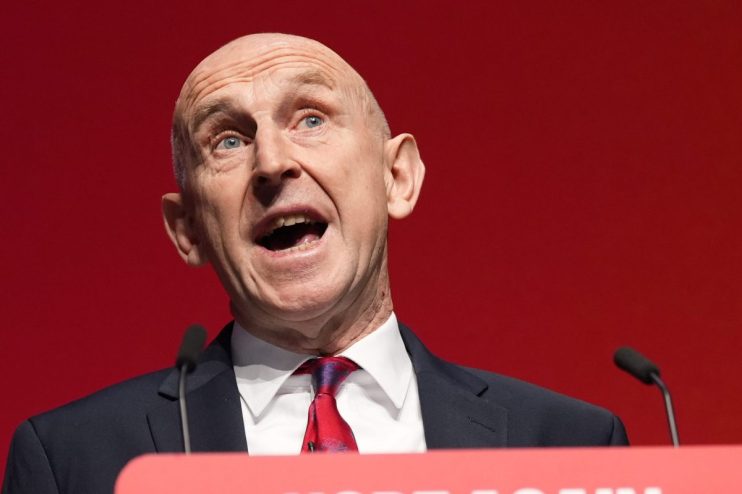 A bald man in a dark suit and red tie speaks passionately at a podium against a red background.