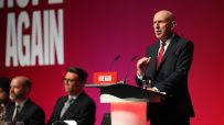 Bald man in suit and red tie speaking at a podium with HOPE AGAIN sign, red background, audience blurred