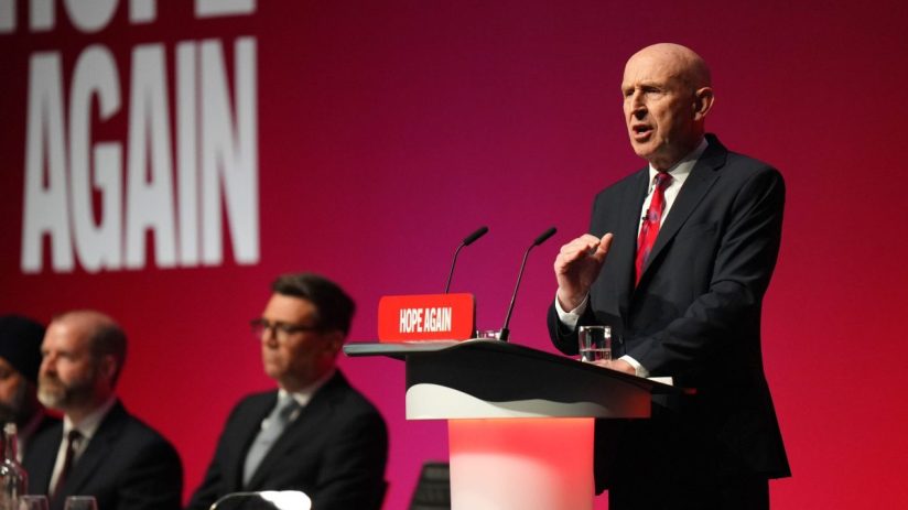Bald man in suit and red tie speaking at a podium with HOPE AGAIN sign, red background, audience blurred