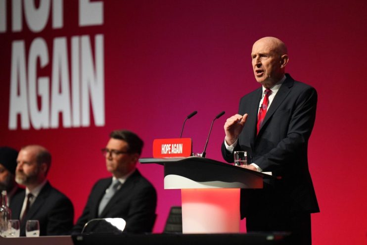 Bald man in suit and red tie speaking at a podium with HOPE AGAIN sign, red background, audience blurred