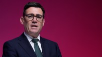 Andy Burnham, Mayor of Greater Manchester, speaking at a podium against a red backdrop.