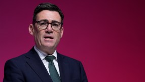 Andy Burnham, Mayor of Greater Manchester, speaking at a podium against a red backdrop.
