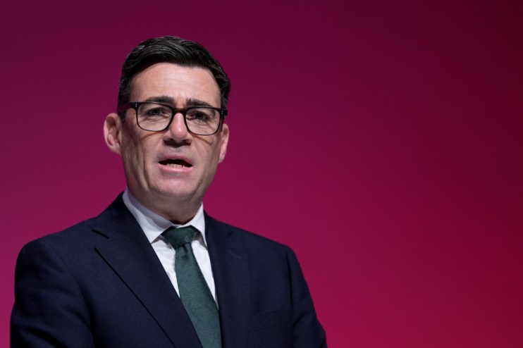 Andy Burnham, Mayor of Greater Manchester, speaking at a podium against a red backdrop.