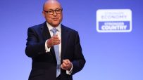 Man in suit and glasses speaking at a podium with a Stronger Economy, Stronger Country sign.
