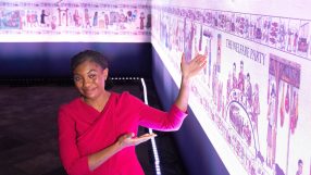 A smiling woman in a red dress gestures towards a long, illuminated historical tapestry with various scenes.
