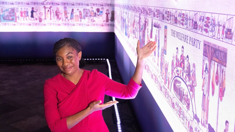 A smiling woman in a red dress gestures towards a long, illuminated historical tapestry with various scenes.