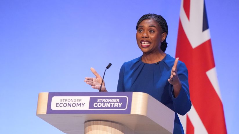 Kemi Badenoch speaking at a podium with Stronger Economy, Stronger Country text and a Union Jack flag.