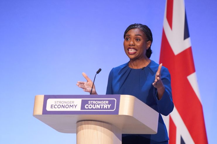 Kemi Badenoch speaking at a podium with Stronger Economy, Stronger Country text and a Union Jack flag.