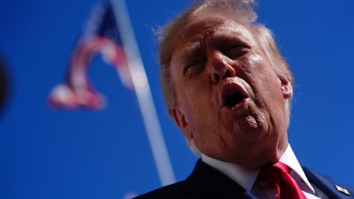 Donald Trump speaking outdoors, wearing a suit and red tie, with an American flag in the blurred background.