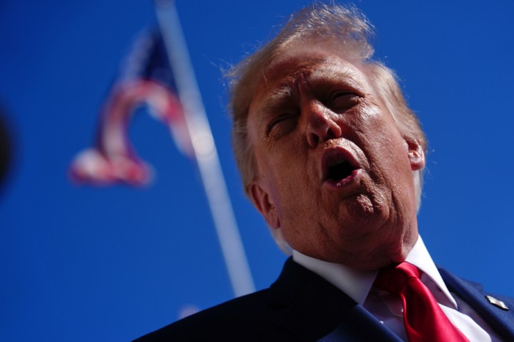 Donald Trump speaking outdoors, wearing a suit and red tie, with an American flag in the blurred background.