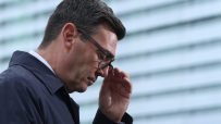 A man in a dark jacket and glasses adjusts his eyewear, looking down with a thoughtful expression.