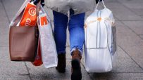 Person walking away with multiple shopping bags, including a brown handbag and bags from Guineys and Sam.