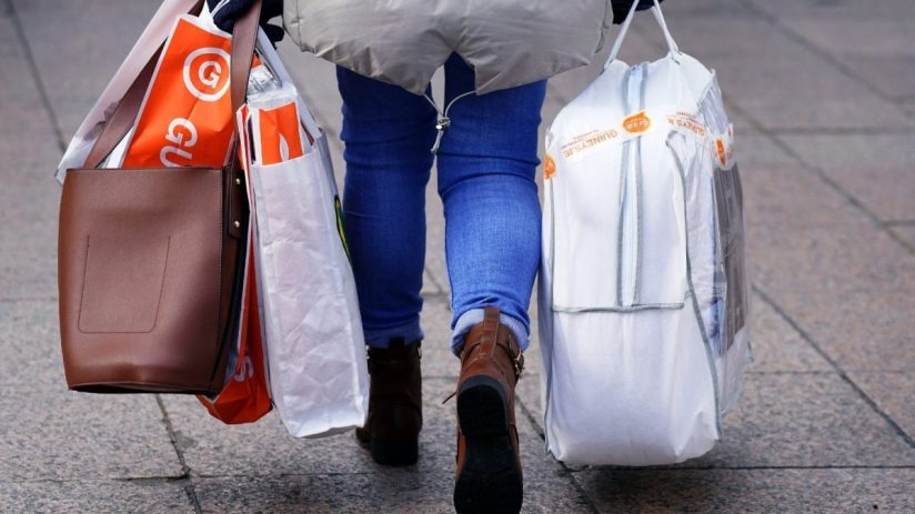 Person walking away with multiple shopping bags, including a brown handbag and bags from Guineys and Sam.