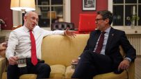Two men in suits, one with a red tie and the other with a dark red tie, sit on a yellow sofa and converse.