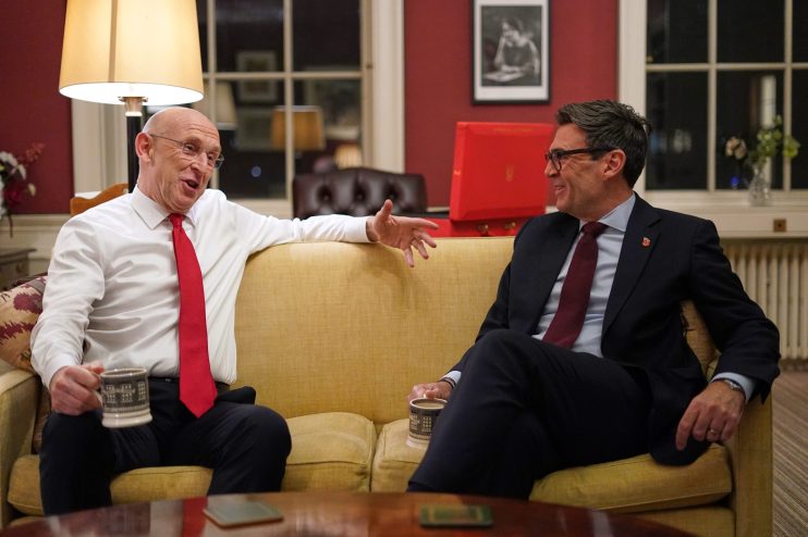 Two men in suits, one with a red tie and the other with a dark red tie, sit on a yellow sofa and converse.