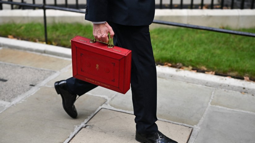 A person in a dark suit walking, carrying the red Chancellor of the Exchequer budget box with the royal crest.