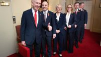 Group of six officials in suits, wearing poppies, one holding a red briefcase, standing in a line on a red carpet.