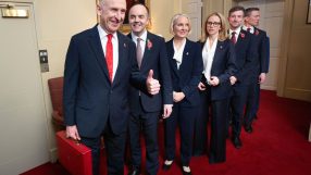 Group of six officials in suits, wearing poppies, one holding a red briefcase, standing in a line on a red carpet.