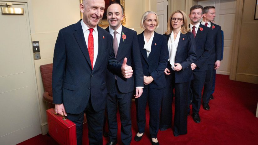 Group of six officials in suits, wearing poppies, one holding a red briefcase, standing in a line on a red carpet.