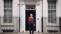 A man in a suit holding a red budget box in front of a black door marked 11