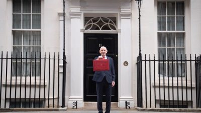 A man in a suit holding a red budget box in front of a black door marked 11