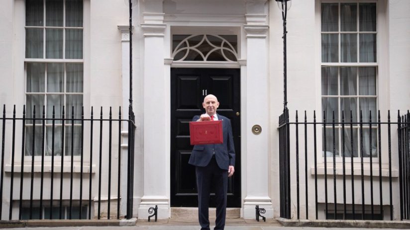 A man in a suit holding a red budget box in front of a black door marked 11
