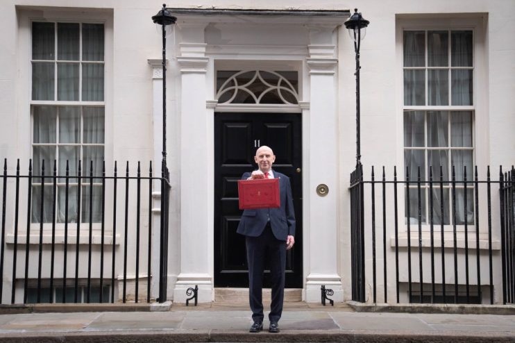 A man in a suit holding a red budget box in front of a black door marked 11