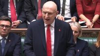 Bald man in glasses speaking from behind a podium, wearing a suit and red tie with a poppy pin.