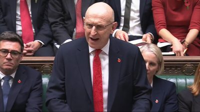 Bald man in glasses speaking from behind a podium, wearing a suit and red tie with a poppy pin.