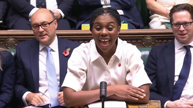 Kemi Badenoch speaks at a microphone in Parliament, flanked by two men in suits, one wearing a poppy.