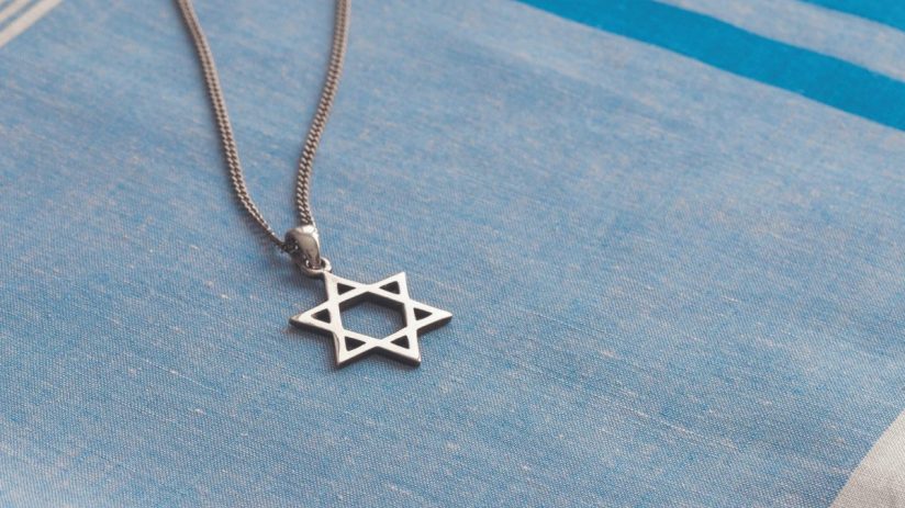 Silver Star of David pendant on a textured blue and white striped fabric, likely a tallit.
