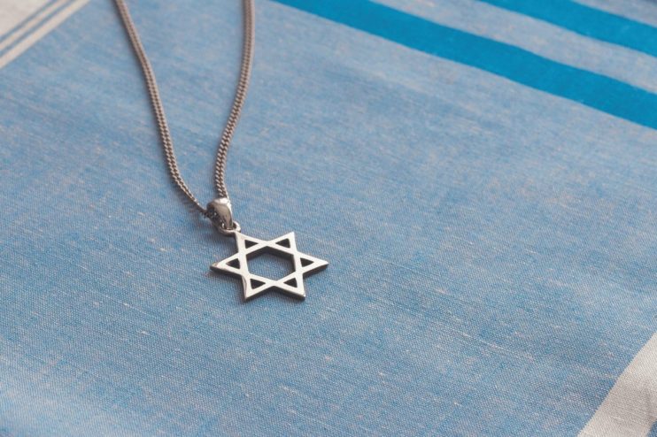 Silver Star of David pendant on a textured blue and white striped fabric, likely a tallit.