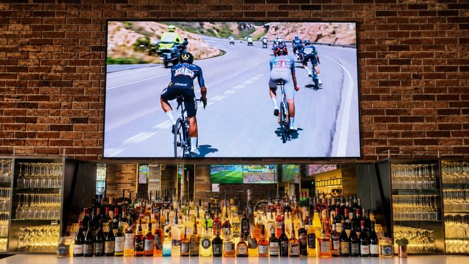 Sports bar interior with a large screen showing a cycling race above a well-stocked liquor bar and brick wall.