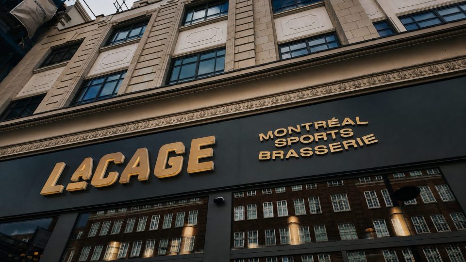 Facade of Montréal Sports Brasserie, Lacage, with gold lettering and reflections of a brick building.