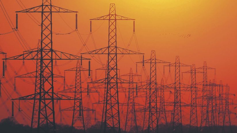 Multiple electricity pylons silhouetted against an orange sunset sky, symbolizing energy infrastructure.