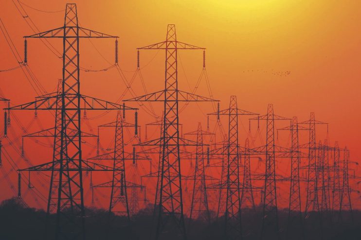 Multiple electricity pylons silhouetted against an orange sunset sky, symbolizing energy infrastructure.