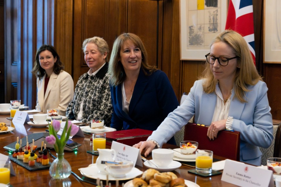 Four women seated at a conference table with refreshments and a British flag in the background.