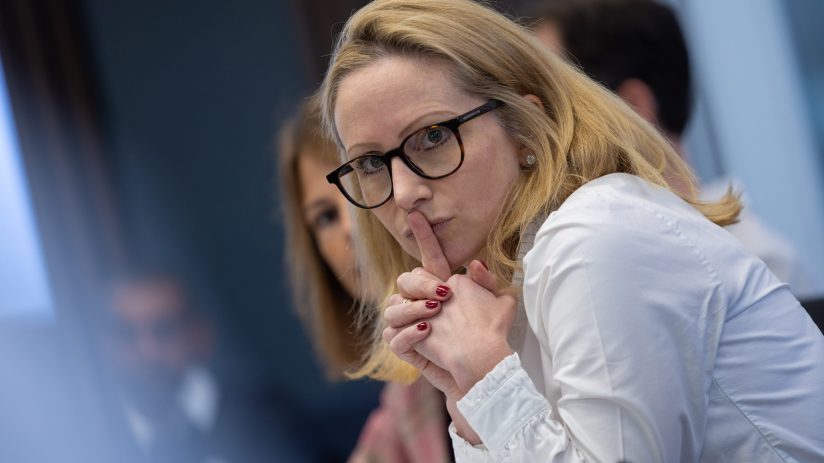 Blonde woman in glasses with finger to lips, a quiet gesture, in a professional meeting setting.
