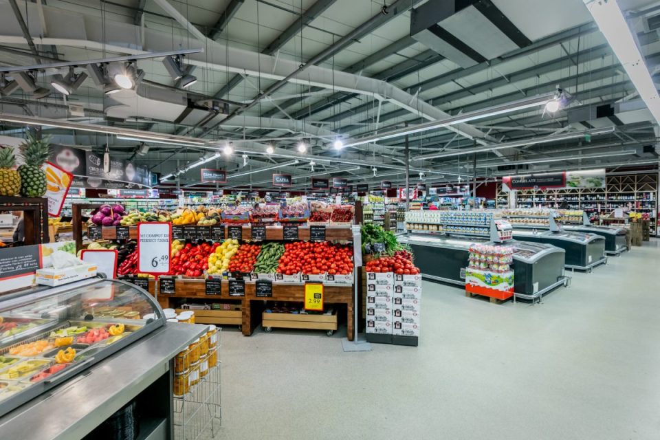 Brightly lit supermarket aisle with fresh produce, pineapples, tomatoes, vegetables, and frozen food displays