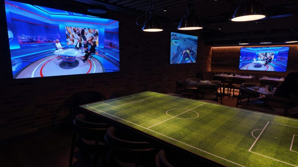 Lounge with a football pitch table, multiple screens showing sports news and Formula 1, and dining tables.