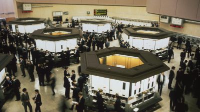 Busy trading floor from the 1980s Big Bang, brokers in suits around hexagonal booths, stock market activity.