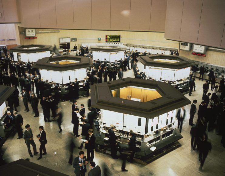 Busy trading floor from the 1980s Big Bang, brokers in suits around hexagonal booths, stock market activity.