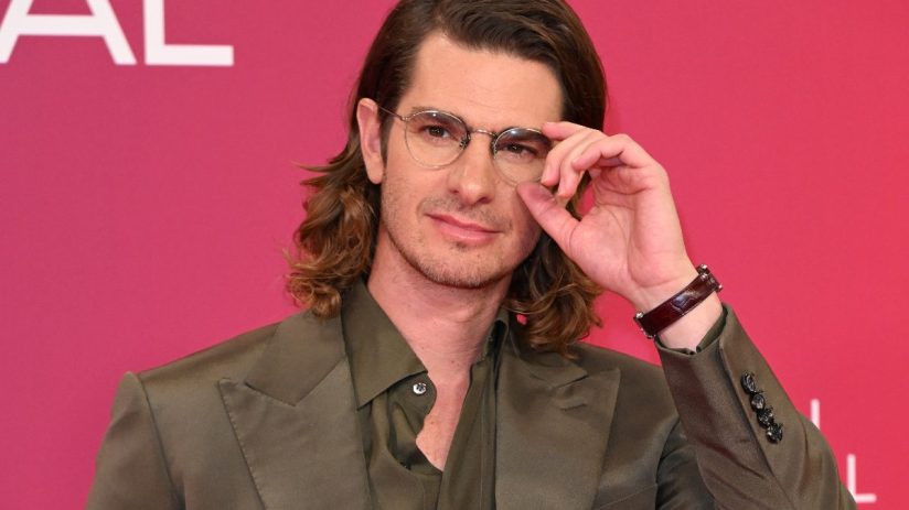 Andrew Garfield in a green suit and glasses, hand adjusting his spectacles, against a pink background for AI film news.
